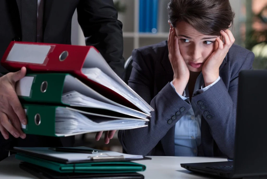 A stressed office employee sits at a desk holding their head while a colleague places a stack of binders on the desk, illustrating heavy workload, workplace pressure, and performance management challenges.