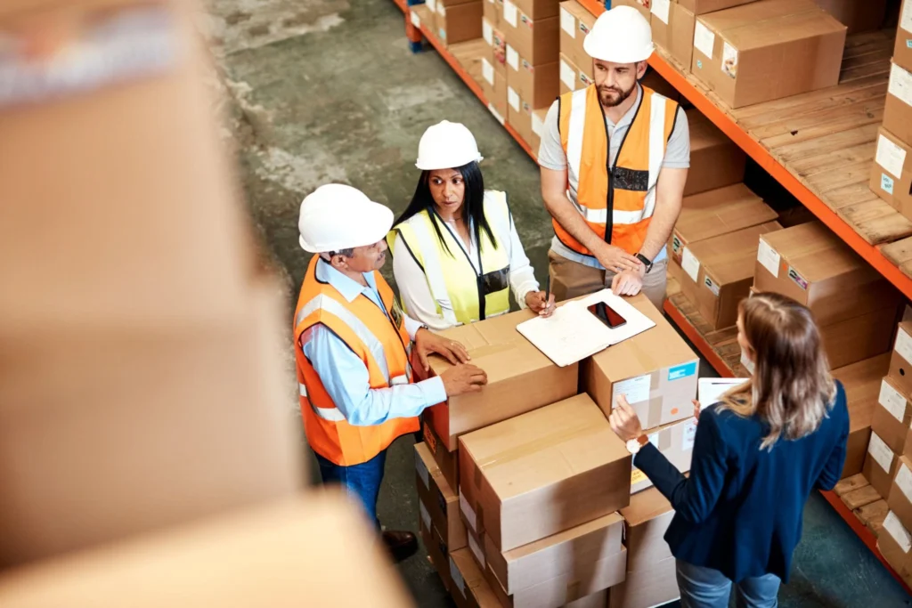Four warehouse employees gathered around stacked packages, reviewing inventory records and coordinating warehouse operations in a large distribution center.