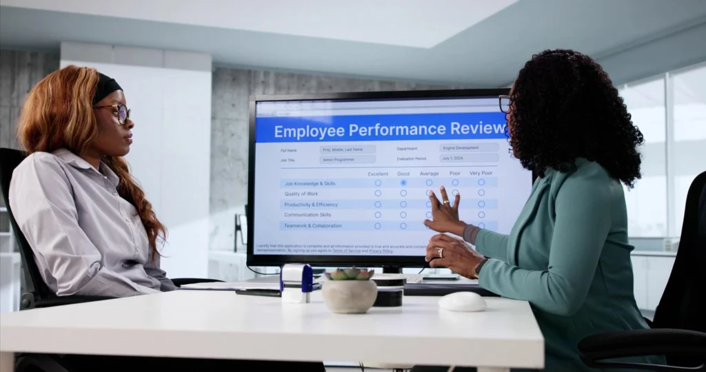 Manager conducting an employee performance review with a team member, discussing evaluation ratings on a digital performance assessment dashboard in a modern office.